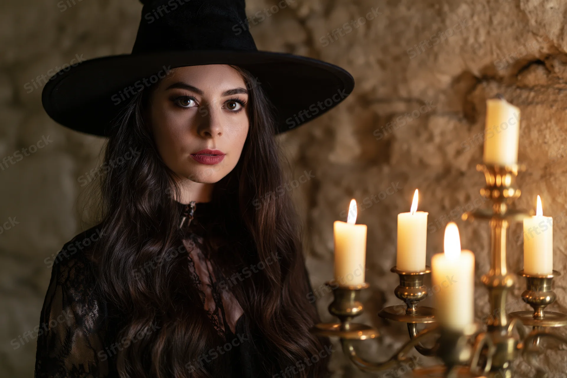 Woman in witch costume with wide-brimmed hat and lace dress standing beside lit candelabra against stone wall
