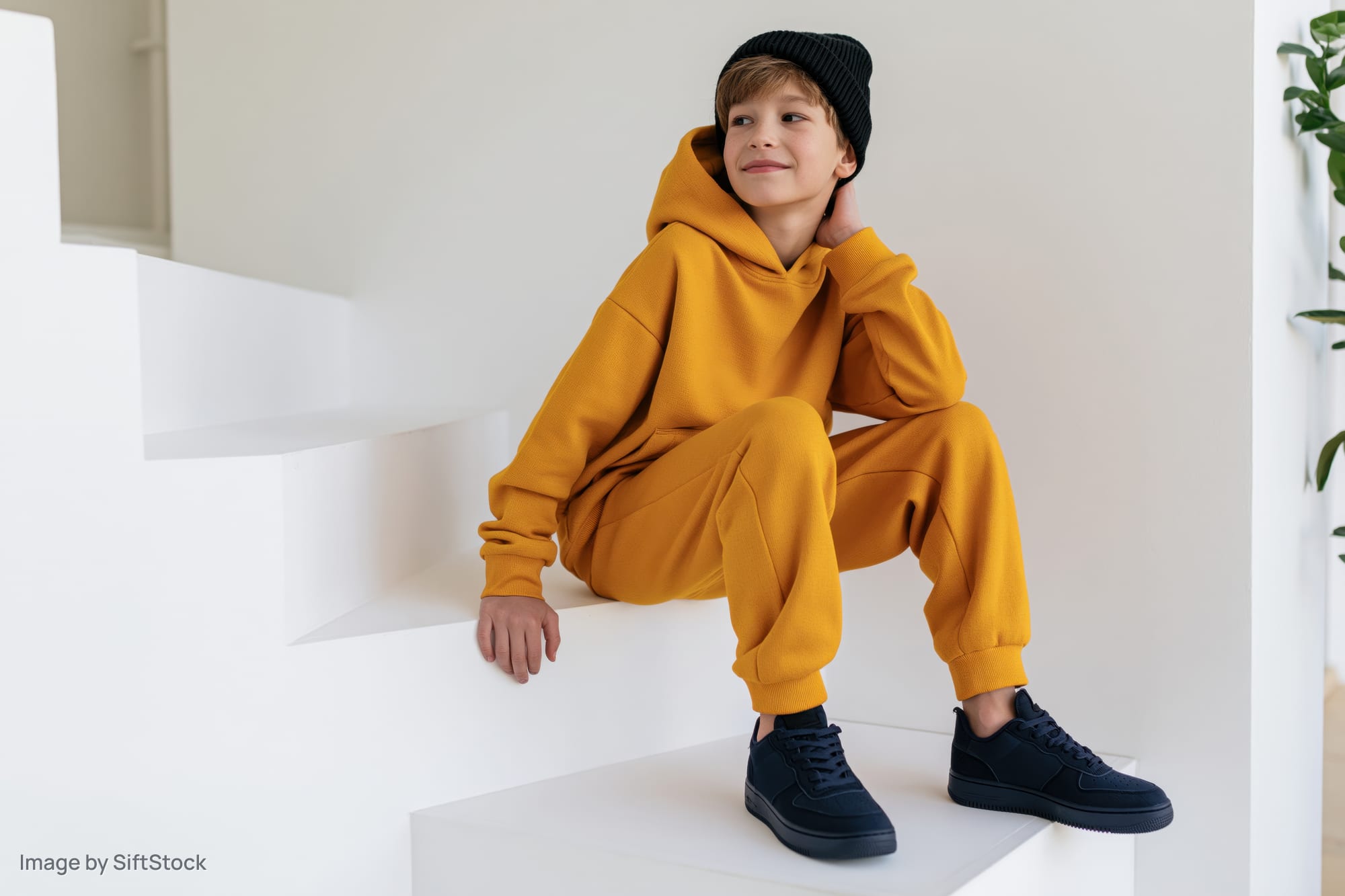 Boy in yellow hoodie and black beanie sitting on white stairs indoors