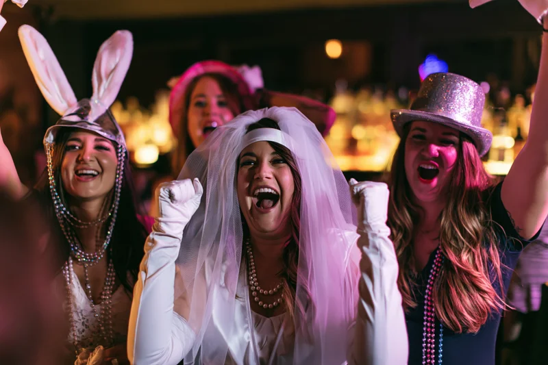 Bride in white veil and gloves celebrating with friends at a bachelorette party in a bar at night