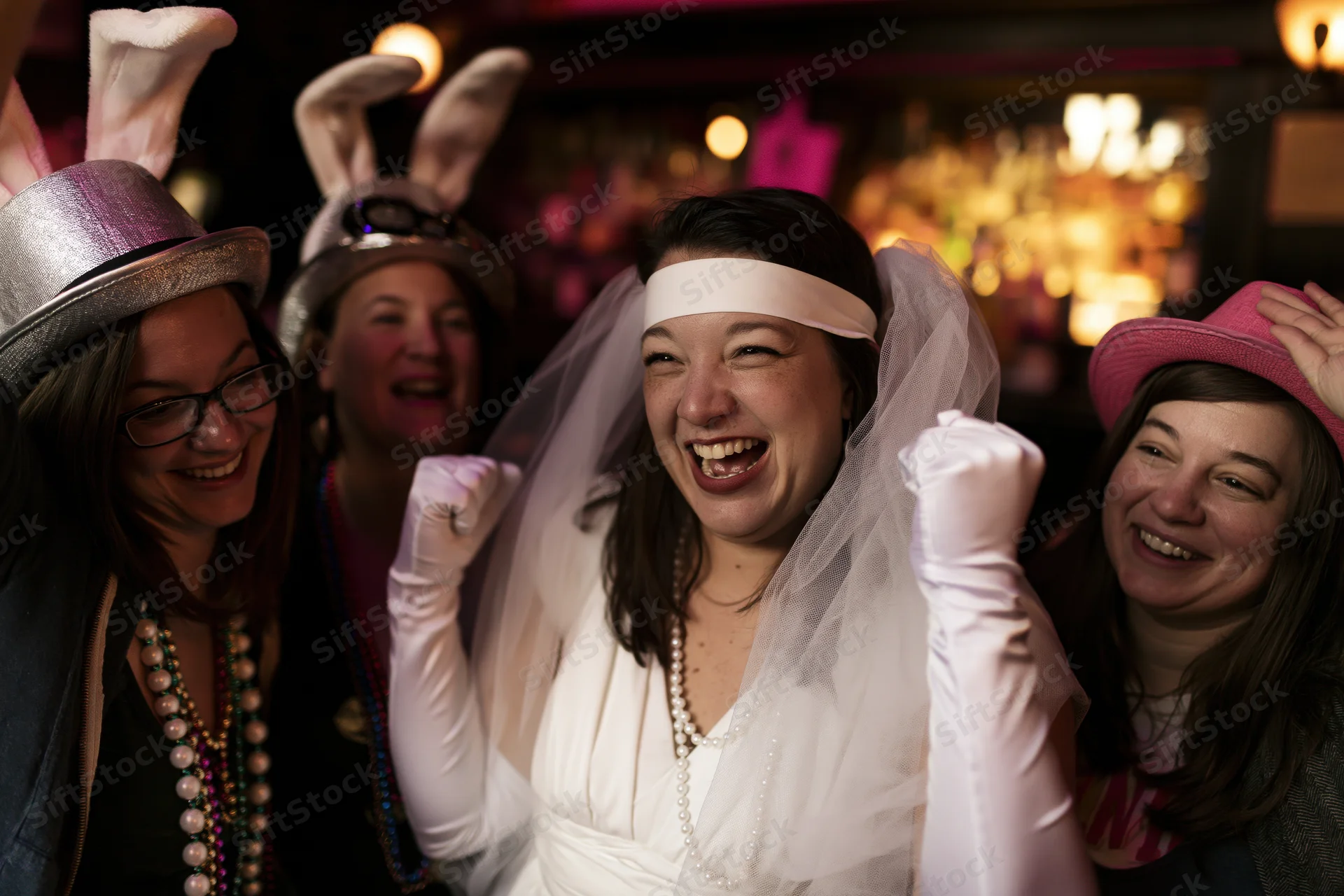 Bride in white veil and long gloves laughing with friends at a bachelorette party in a bar