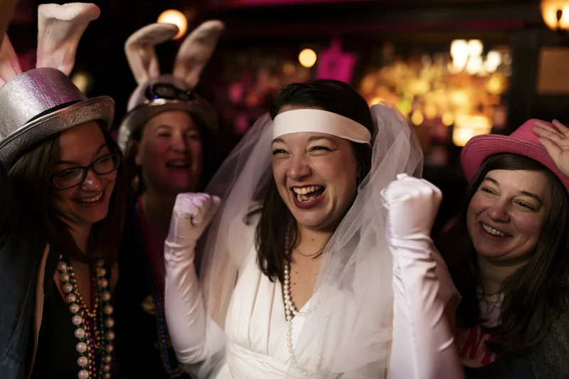 Bride in white veil and long gloves laughing with friends at a bachelorette party in a bar