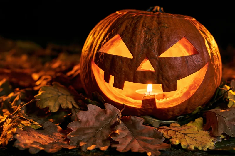 Carved pumpkin with glowing candle face resting among dry autumn leaves at night