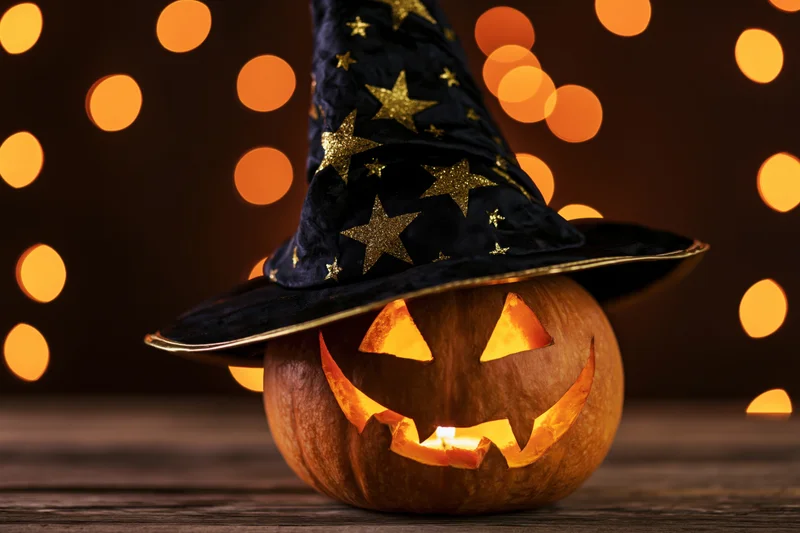 Carved pumpkin with witch hat glowing on wooden table against bokeh lights