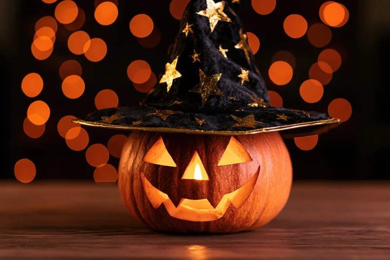 Carved pumpkin with witch hat glowing on wooden table against orange bokeh lights