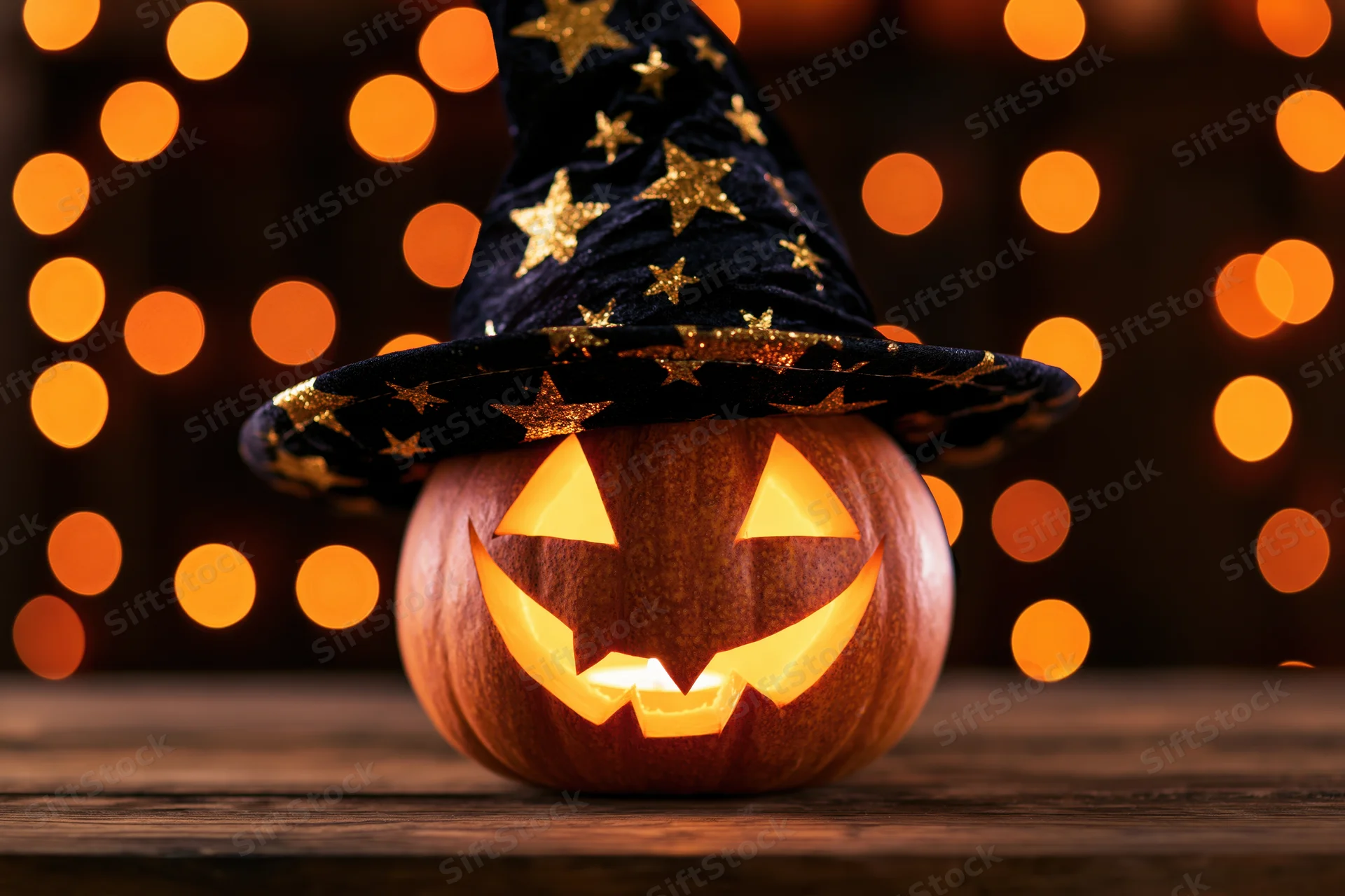 Carved jack-o-lantern with glowing face wearing a black witch hat with gold stars on a wooden table against warm bokeh lights