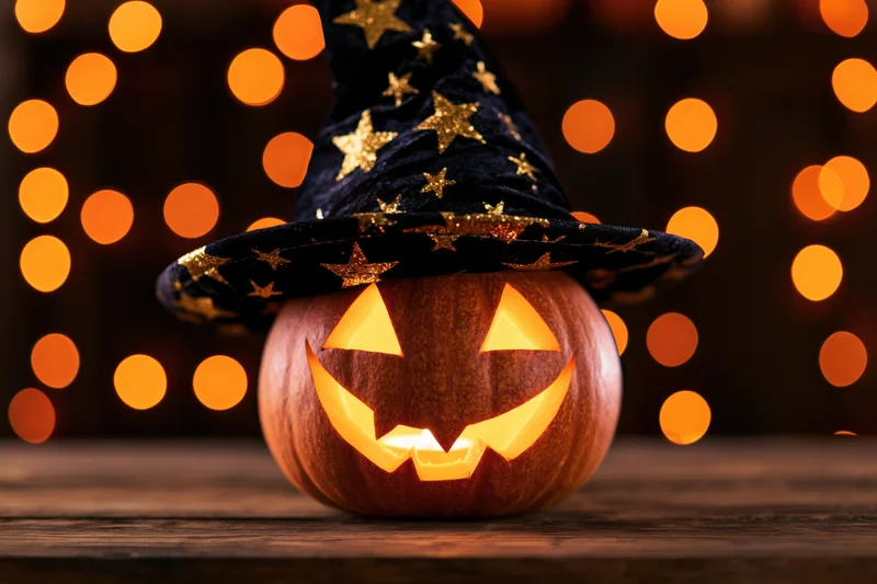 Carved jack-o-lantern with glowing face wearing a black witch hat with gold stars on a wooden table against warm bokeh lights
