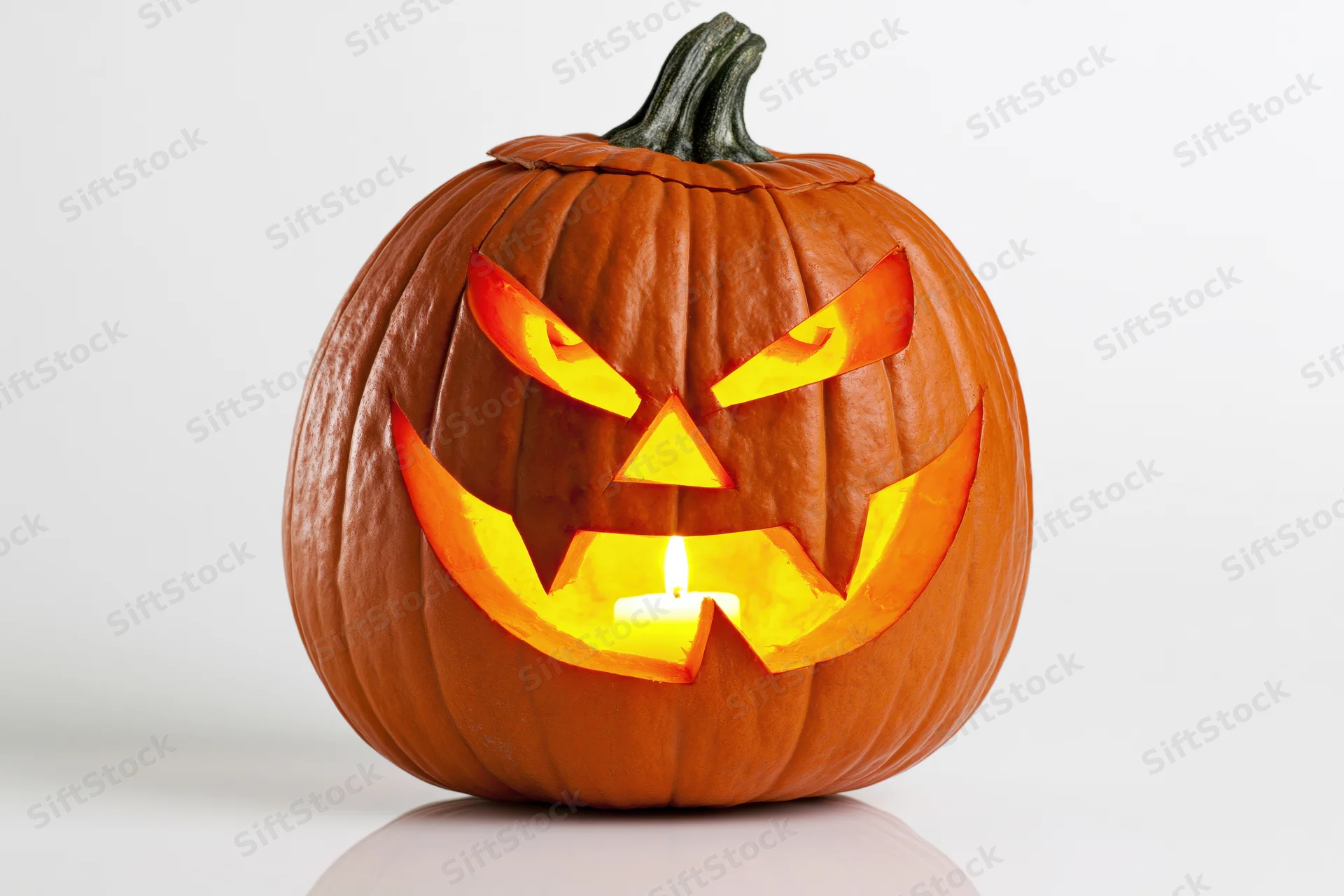Carved pumpkin with angular menacing face illuminated by candle on white surface