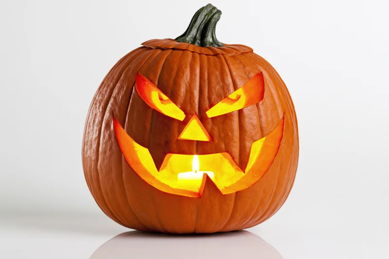 Carved pumpkin with angular menacing face illuminated by candle on white surface
