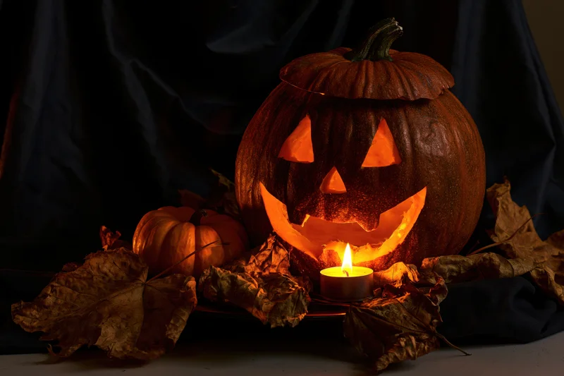Carved pumpkin with glowing candle inside, surrounded by dried leaves on dark cloth