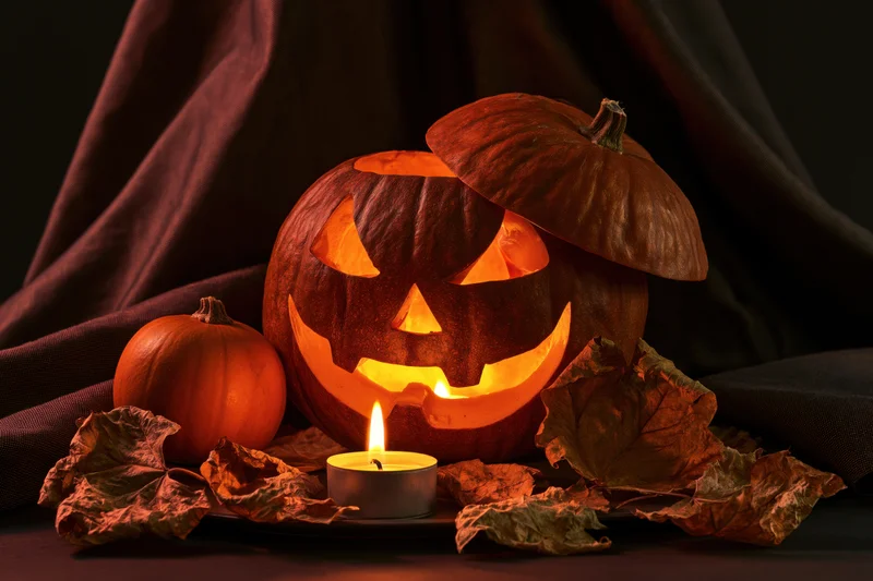 Carved pumpkin with glowing candle inside, surrounded by dried leaves and fabric, for Halloween decoration