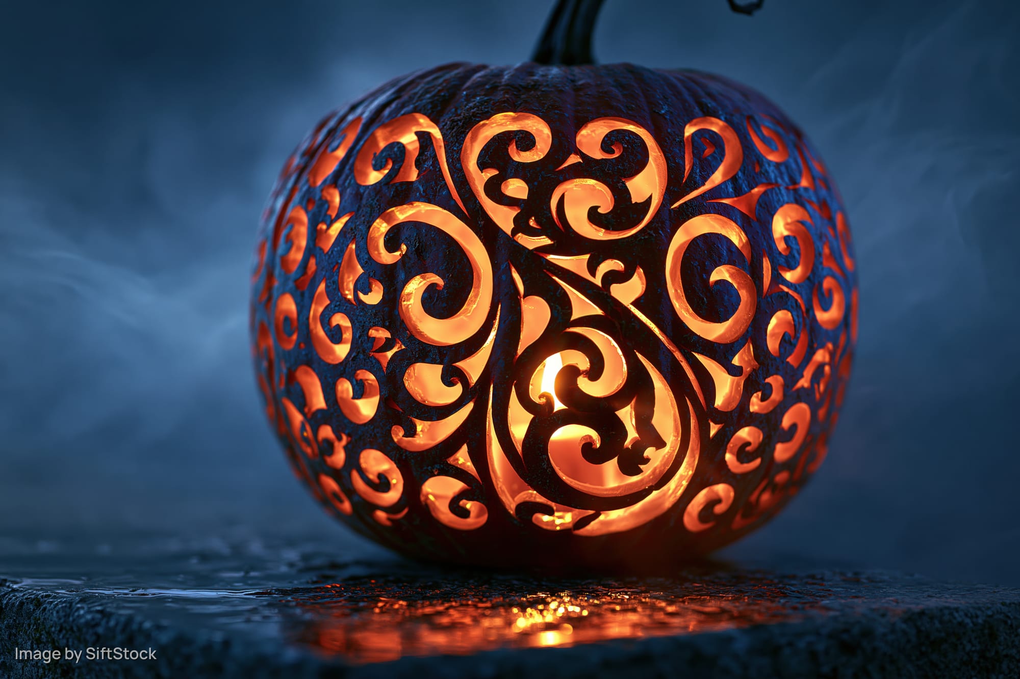 Carved pumpkin with intricate swirling filigree design glowing in the dark on a wet stone surface