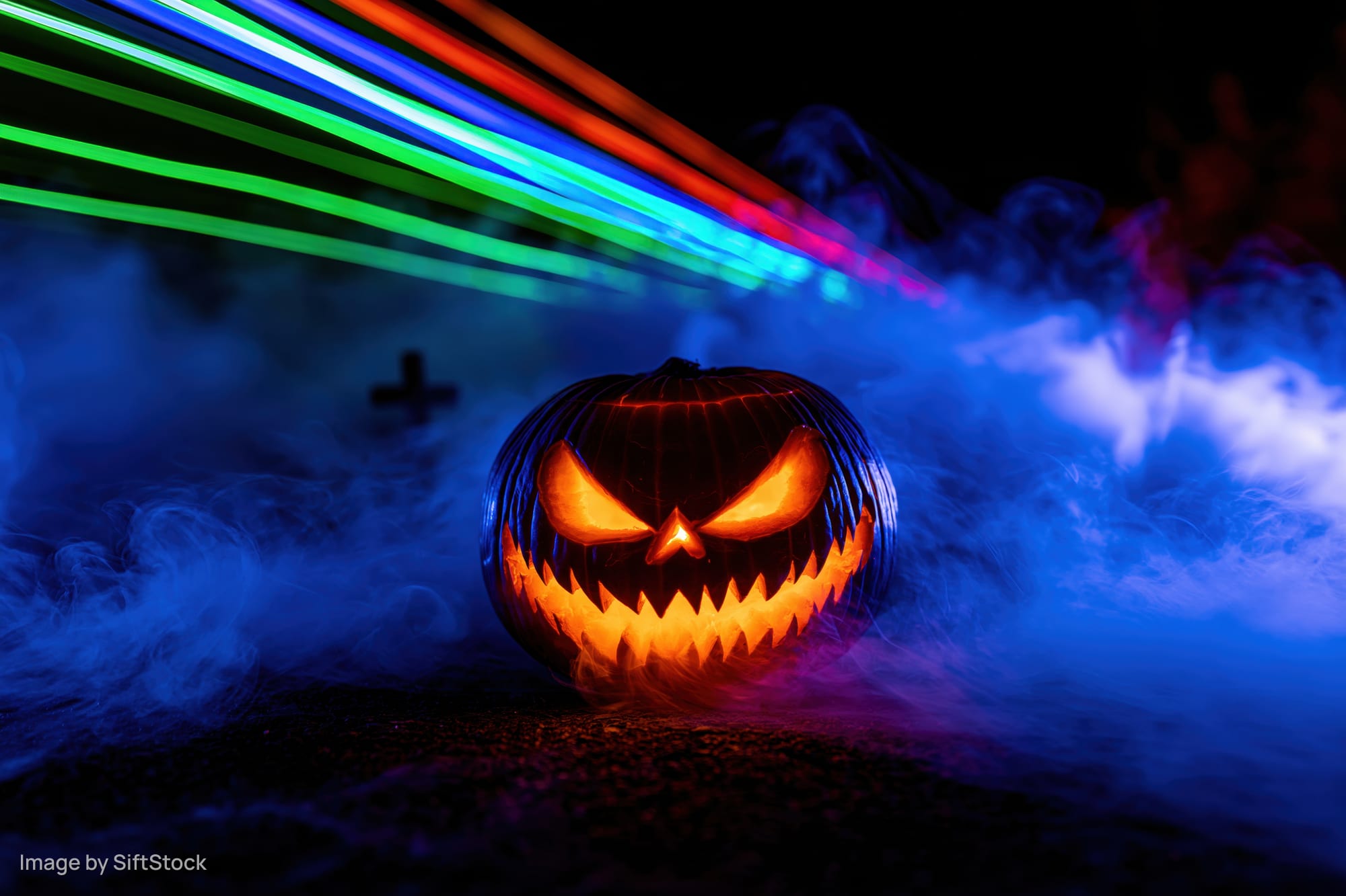 Carved jack-o-lantern with glowing evil grin surrounded by blue smoke and colorful light trails at night
