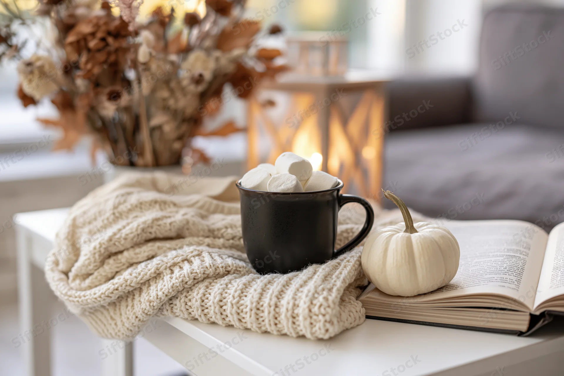 Dark ceramic mug with marshmallows on a knit sweater beside an open book and white pumpkin on a table in a cozy living room