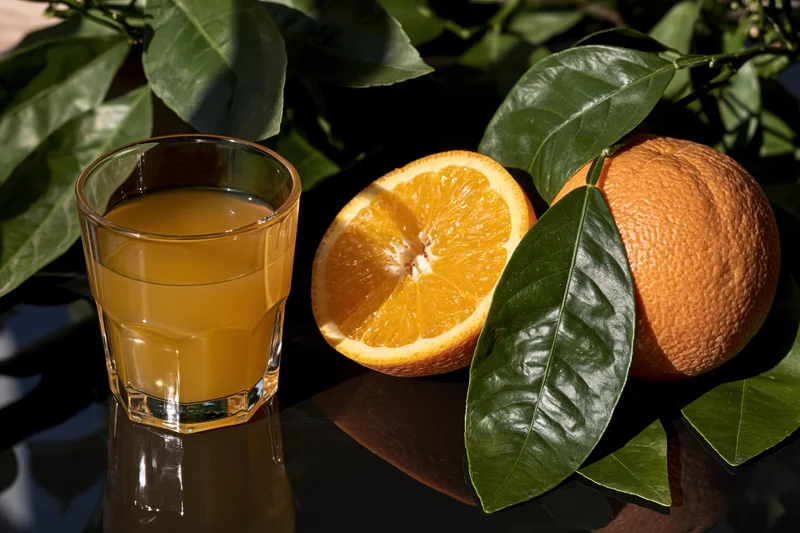 Glass of orange juice beside halved and whole oranges with green leaves on dark reflective surface