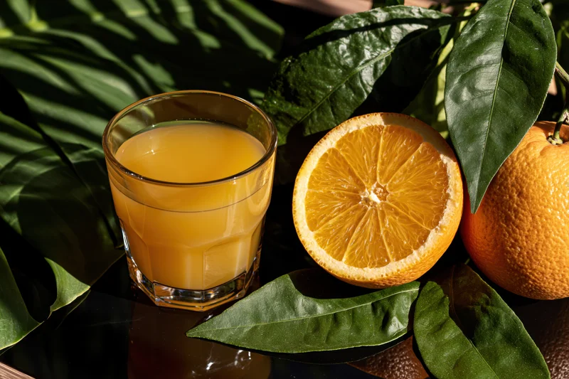 Glass of fresh orange juice beside halved and whole oranges with green leaves on dark reflective surface