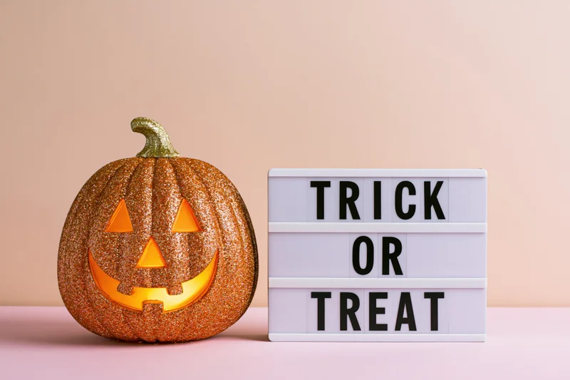 Glittery gold jack-o-lantern with glowing carved face beside a white lightbox sign reading trick or treat on a pastel pink surface