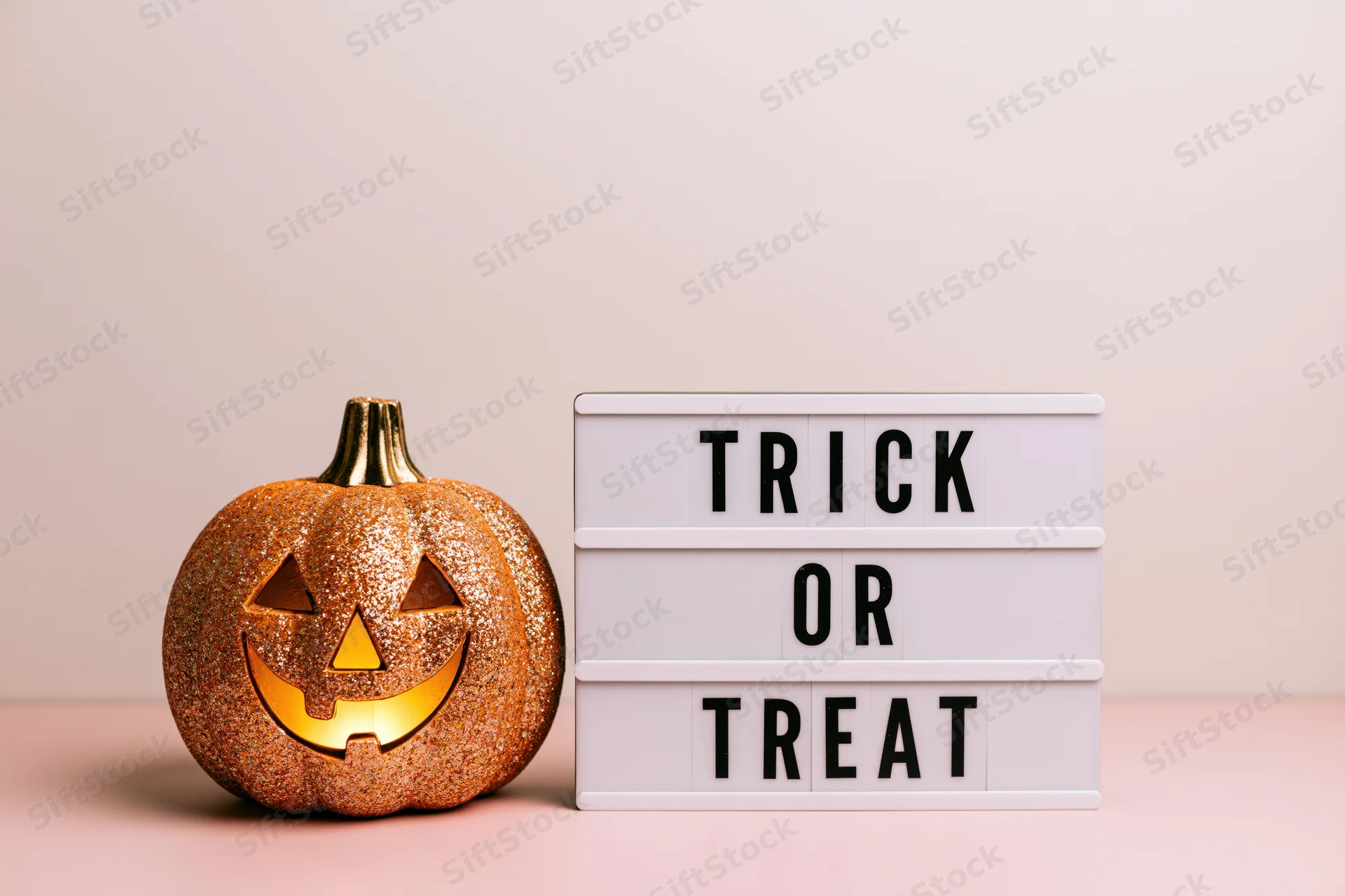 Glittery gold jack-o-lantern with glowing face beside a white lightbox sign reading trick or treat on a pink surface