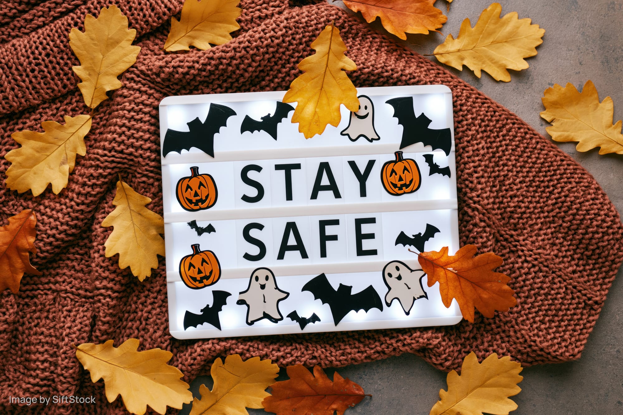 Glowing lightbox with stay safe message and halloween symbols on knitted sweater surrounded by autumn leaves