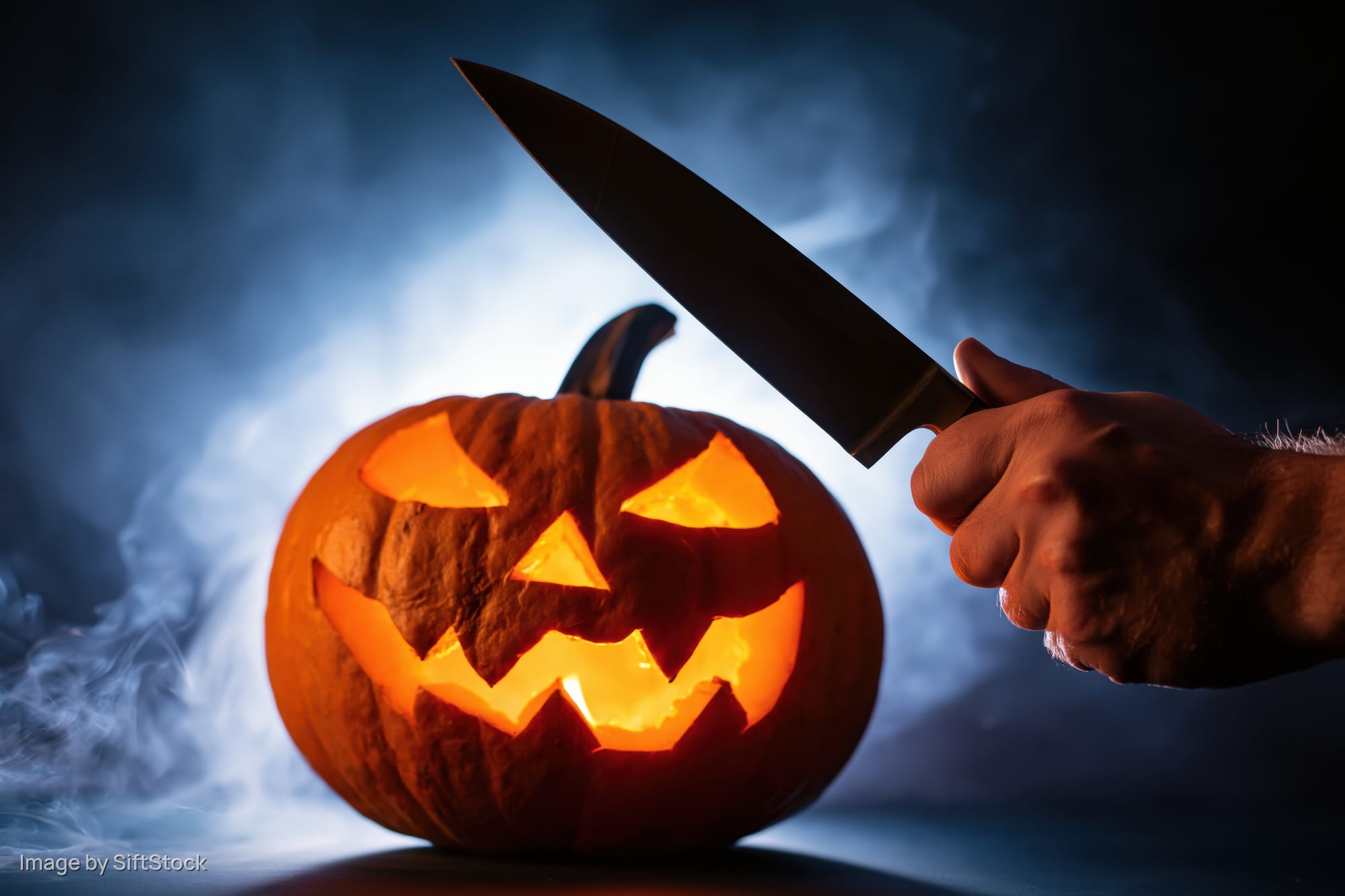 Hand holding knife poised to carve glowing jack-o'-lantern in smoky dark setting