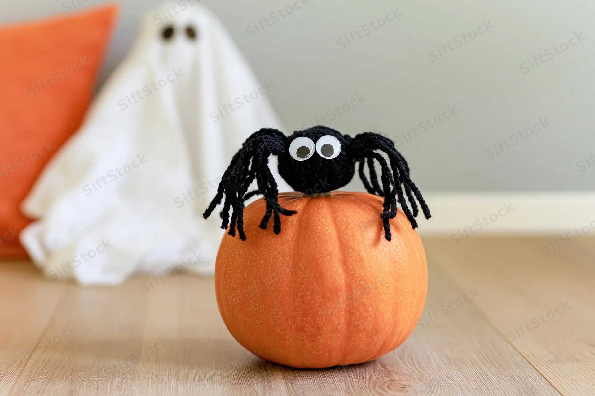 Handmade black yarn spider with googly eyes sitting on orange pumpkin on wooden floor with ghost decoration in background