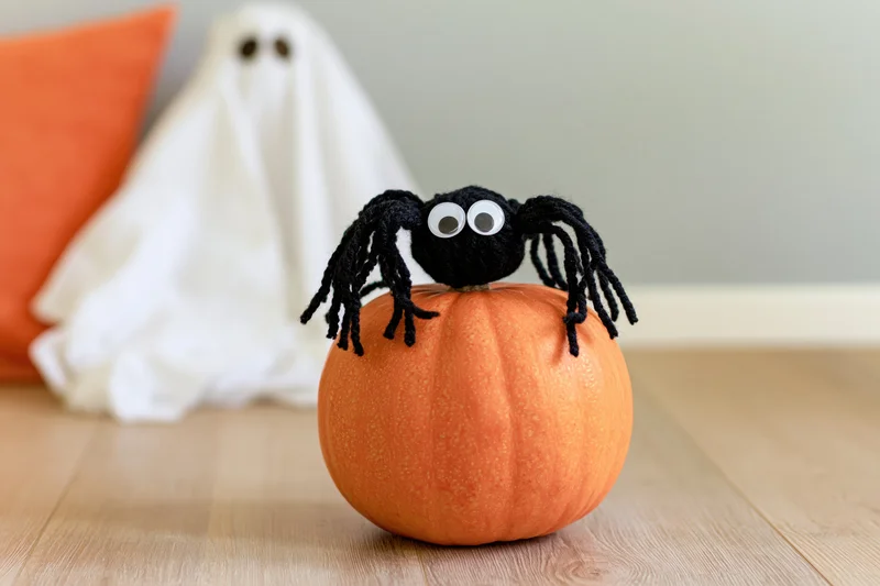 Handmade black yarn spider with googly eyes sitting on orange pumpkin on wooden floor with ghost decoration in background