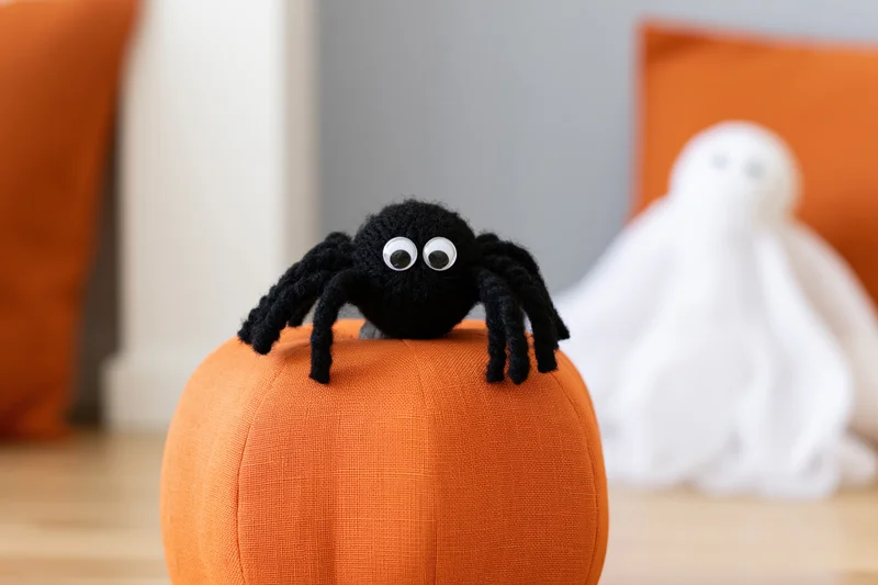 Handmade black yarn spider with googly eyes sitting on orange fabric pumpkin with ghost decoration in background