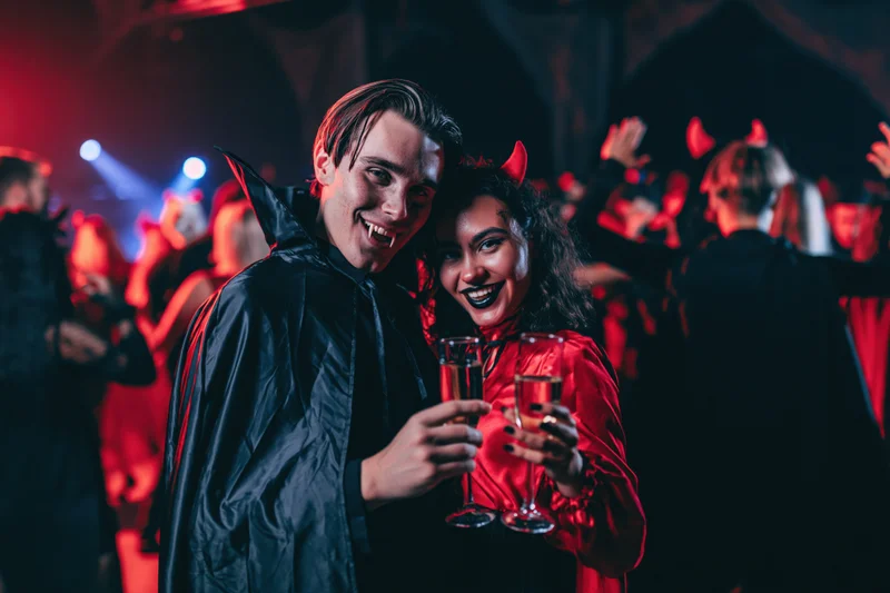 Couple in vampire and devil costumes smiling while holding wine glasses at a halloween party