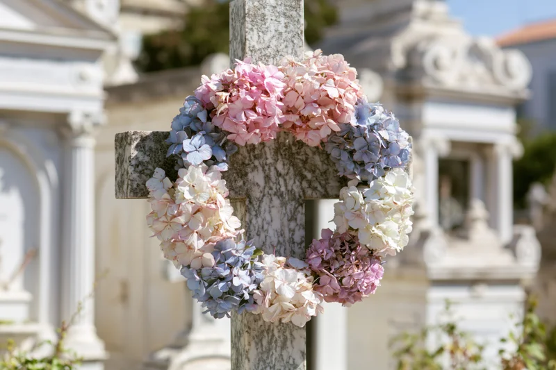 A marble cross in a cemetery adorned with a pink blue and white hydrangea wreath