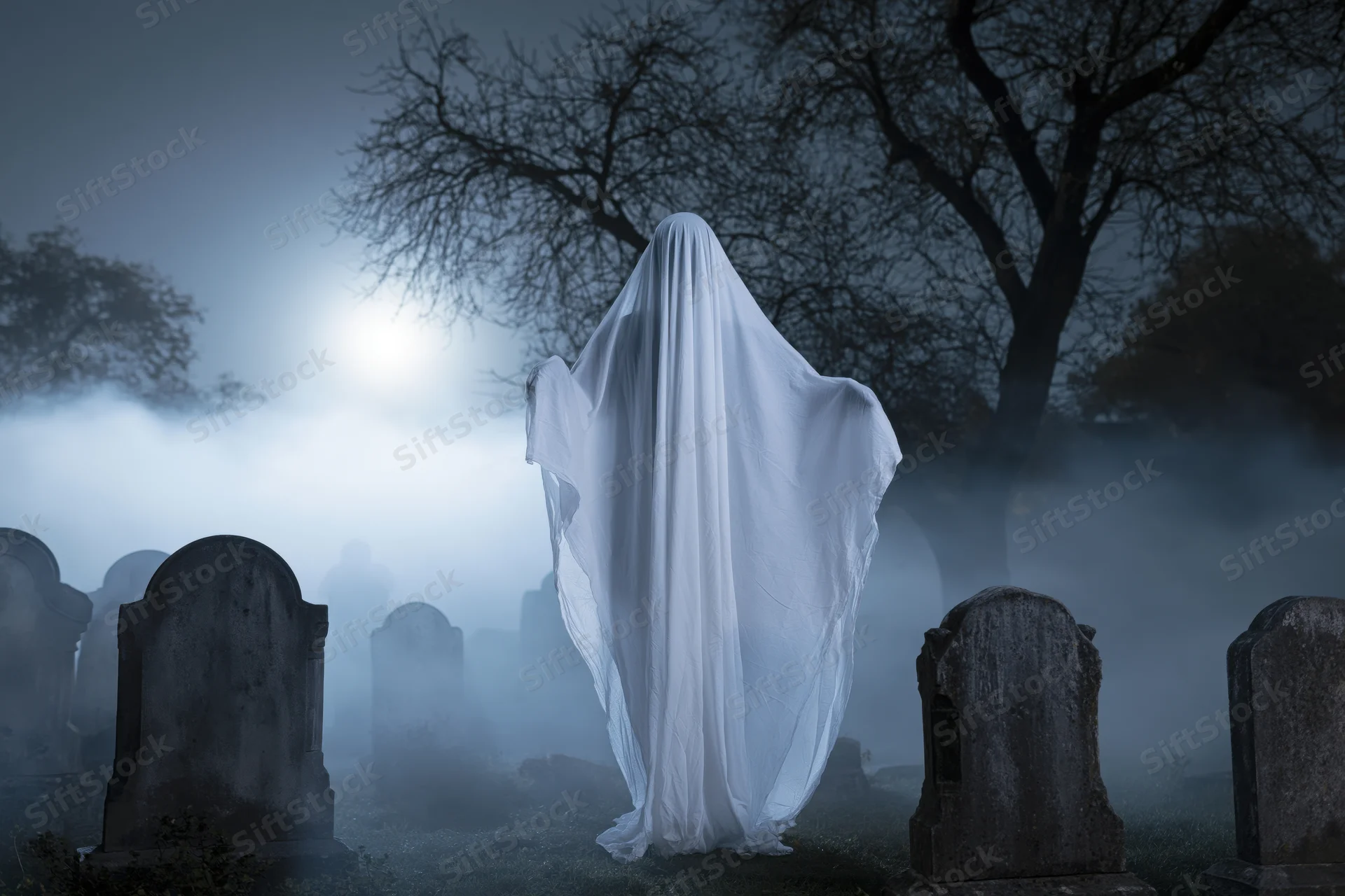 Ghost in flowing white sheet costume standing among tombstones in foggy cemetery at night