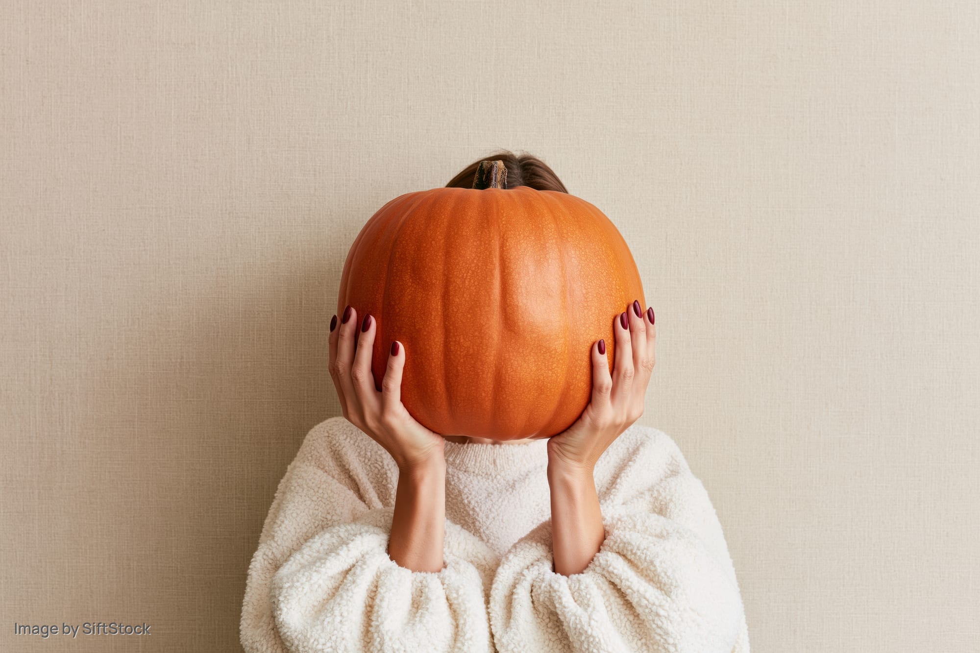 Woman holding large orange pumpkin in front of her face against beige wall