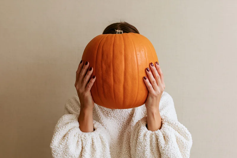 Woman holding large orange pumpkin in front of her face, wearing white fleece sweater against plain wall