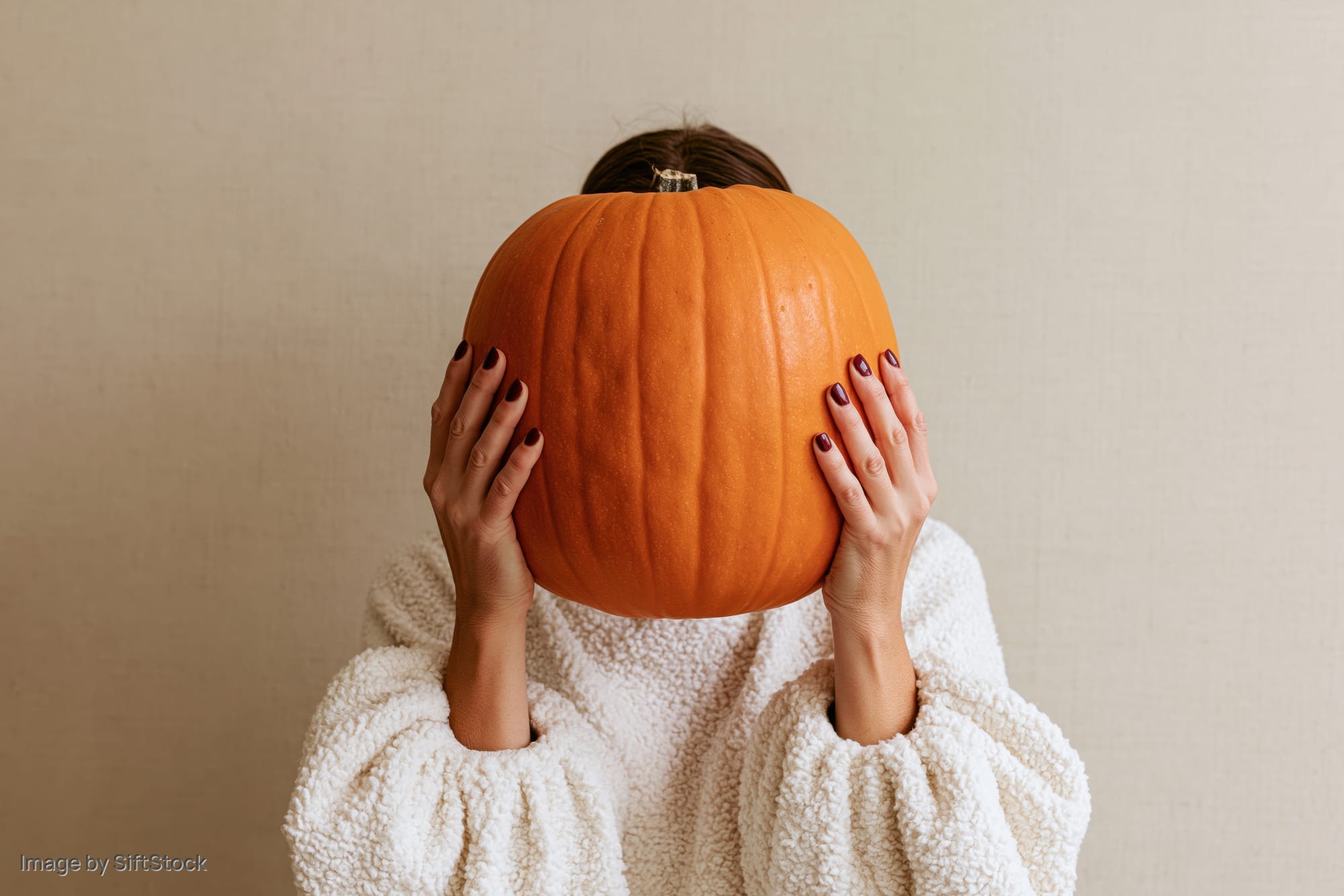 Woman holding large orange pumpkin in front of her face, wearing white fleece sweater against plain wall