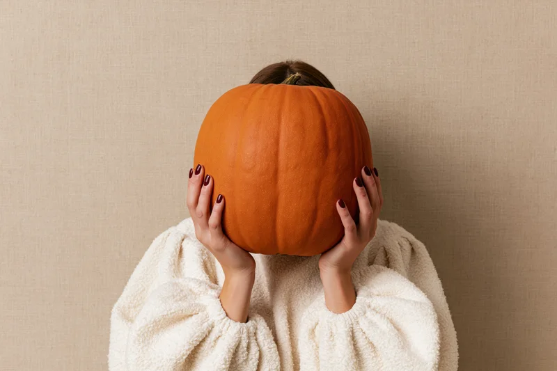 Woman holding large orange pumpkin in front of her face, wearing white fleece sweater against beige wall