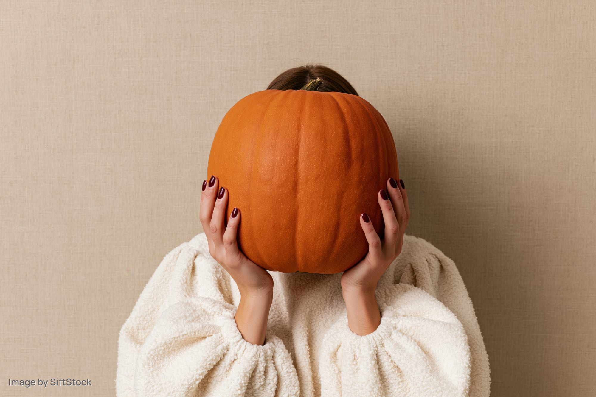 Woman holding large orange pumpkin in front of her face, wearing white fleece sweater against beige wall