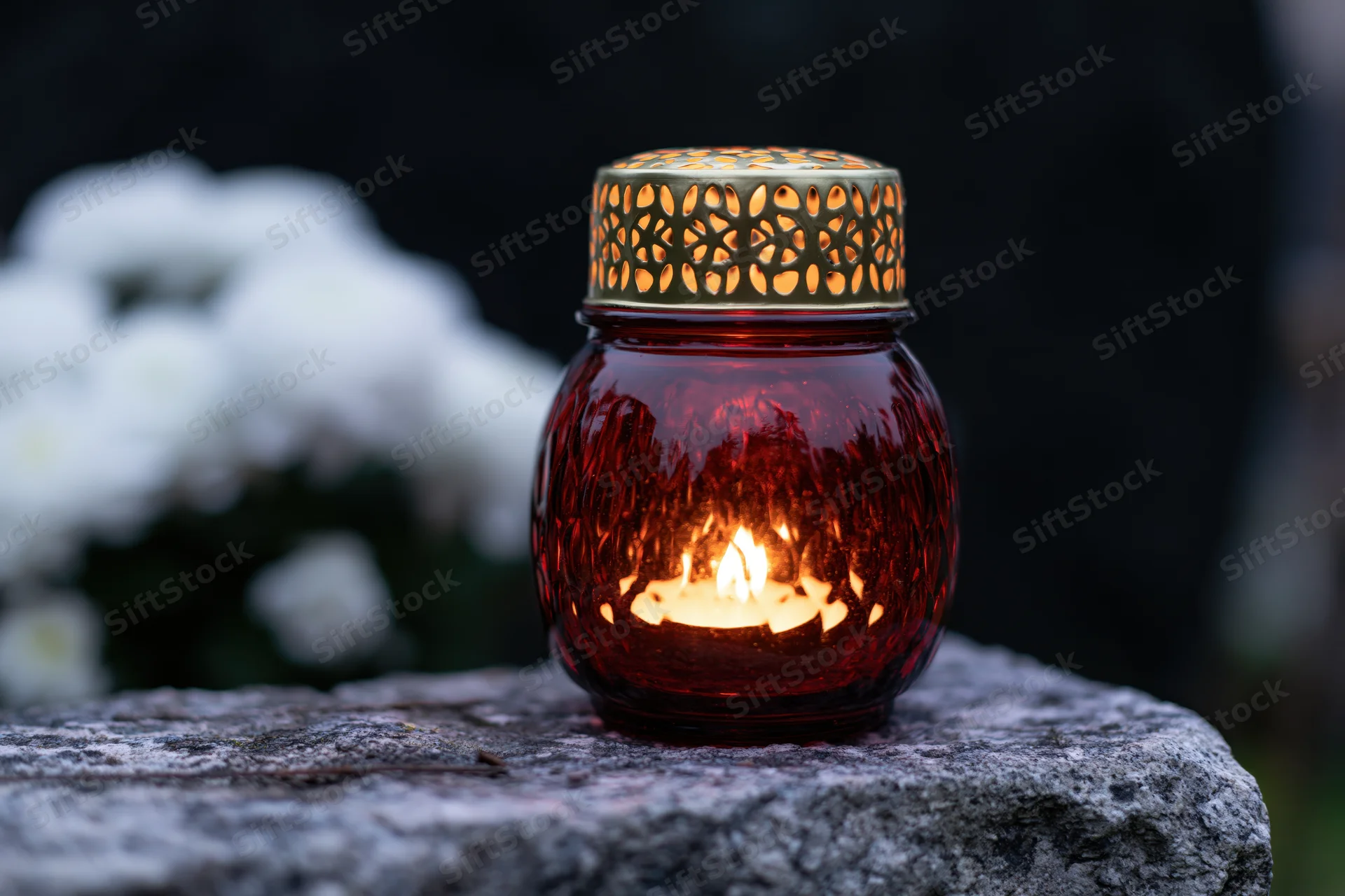 Red glass grave lantern with lit candle and gold perforated lid on stone tombstone, white flowers blurred in background
