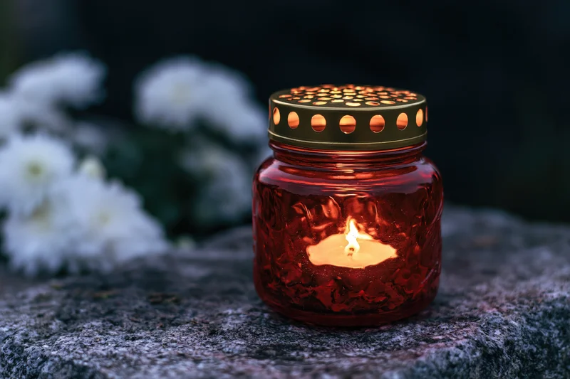Red glass grave lantern with lit candle and gold perforated lid on stone surface near white flowers
