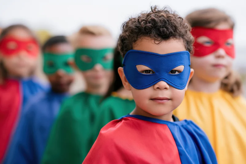 Young children in colorful superhero costumes with masks and capes standing in a row outdoors