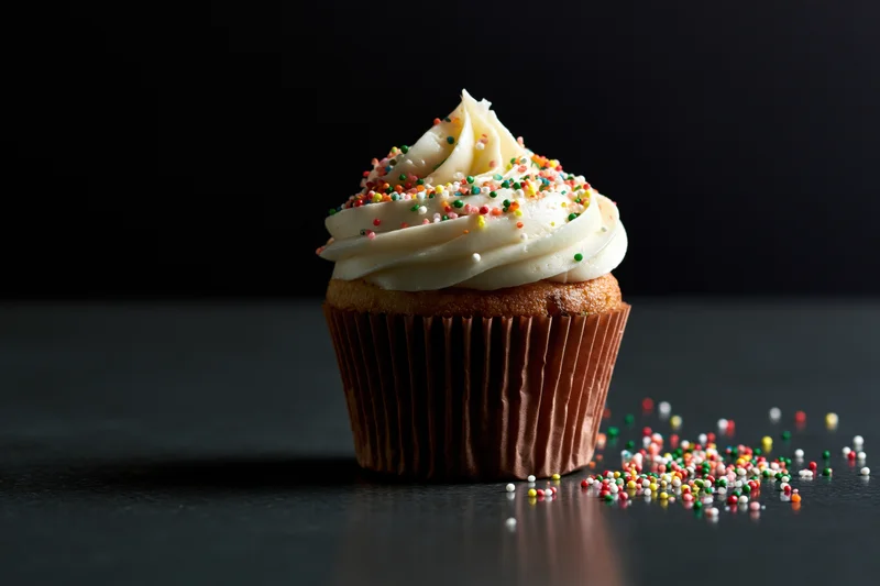 Single cupcake with white frosting and colorful sprinkles on dark surface