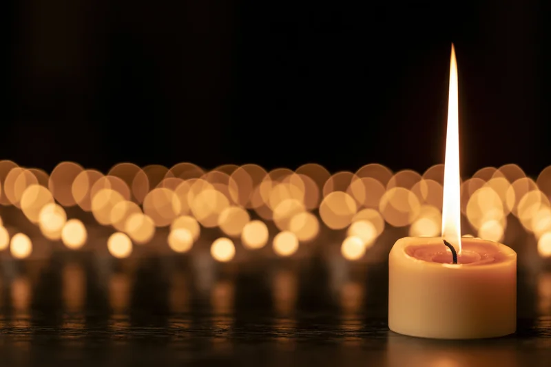 Single burning candle with tall flame in front of blurred golden lights on dark surface