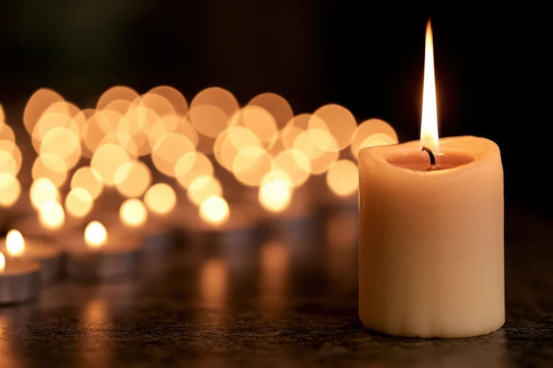Single pillar candle burning with tall flame on dark surface, blurred lights in background