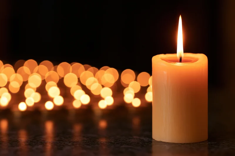 A short pillar candle burning with a tall steady flame on a dark surface, with blurred warm lights in the background