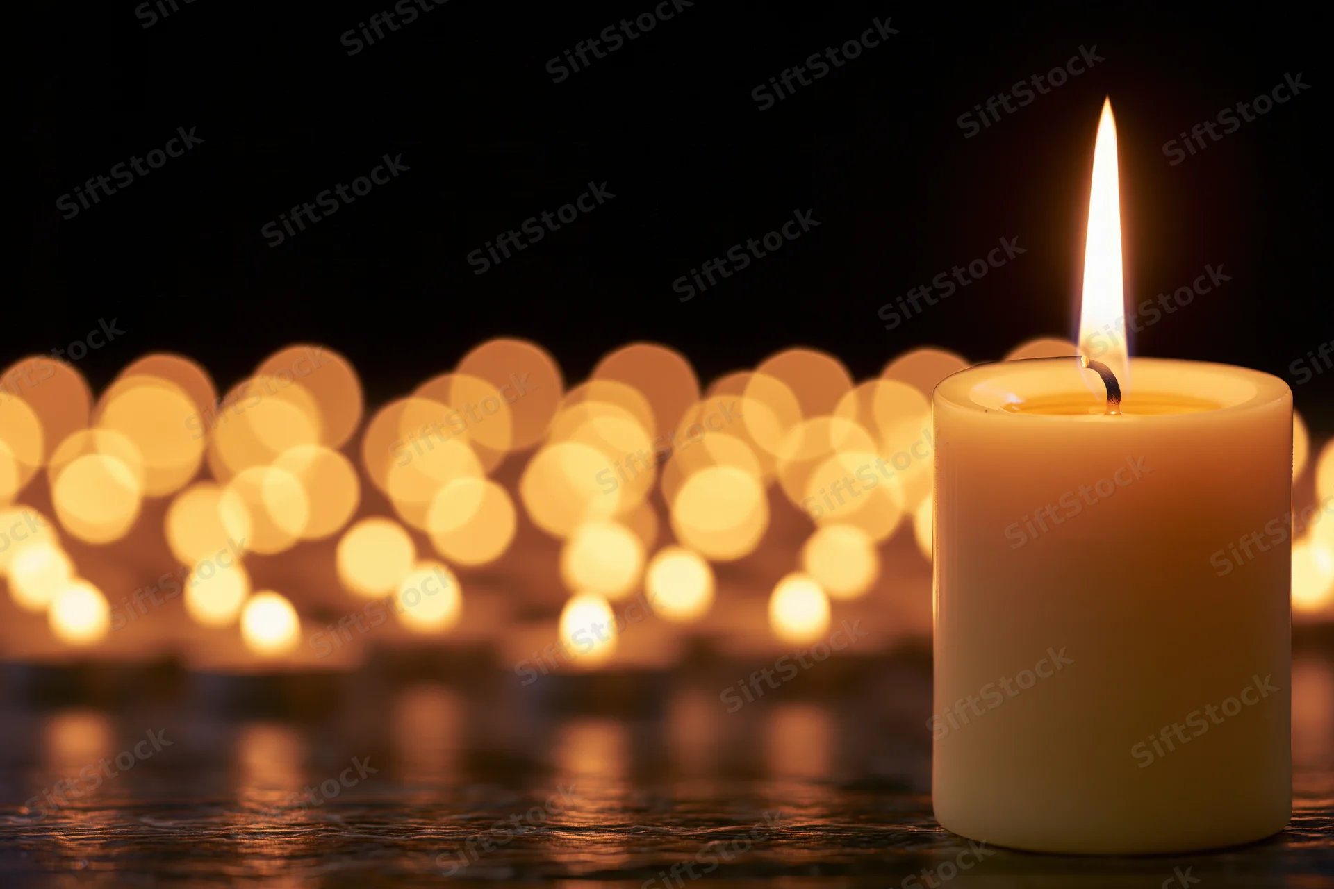 Single lit pillar candle in focus with blurred golden lights in background on dark surface