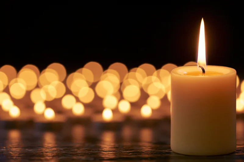 Single lit pillar candle in focus with blurred golden lights in background on dark surface