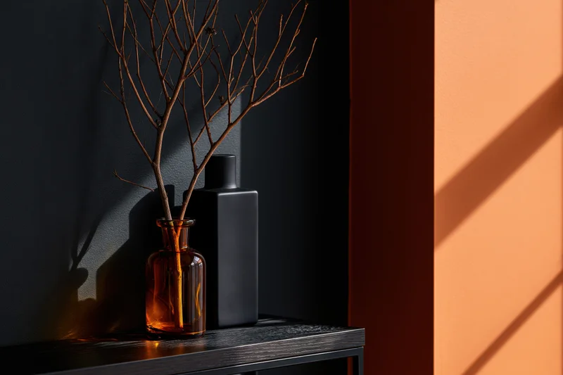 Amber glass vase with dried twig on dark shelf beside orange wall in sunlight