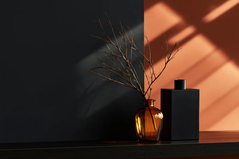 Amber glass vase with dried twig beside black bottle on dark shelf against two-toned wall with sunlight shadows