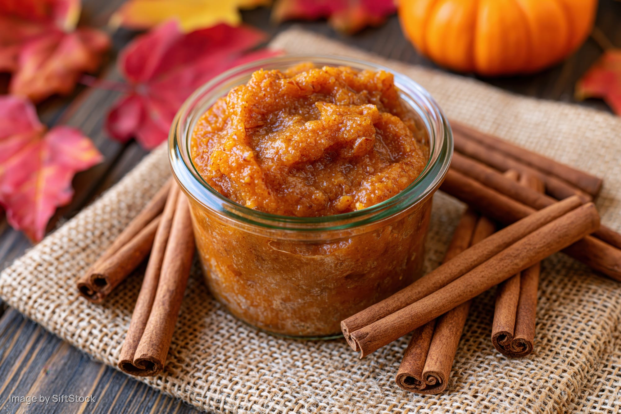 Chunky pumpkin puree in a glass jar surrounded by cinnamon sticks on burlap with autumn leaves and a small pumpkin in the background