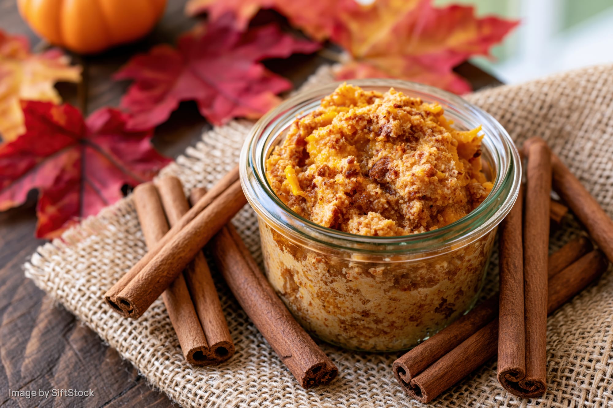 Chunky pumpkin scrub in a glass jar surrounded by cinnamon sticks and autumn leaves on burlap cloth