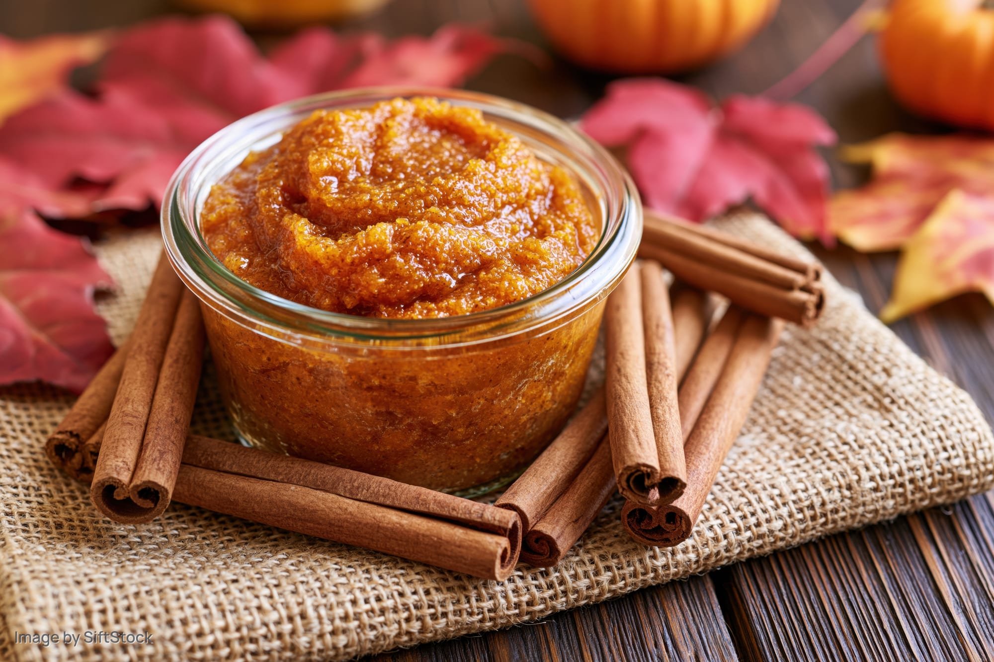 Chunky pumpkin scrub in a glass jar surrounded by cinnamon sticks and autumn leaves on a wooden table
