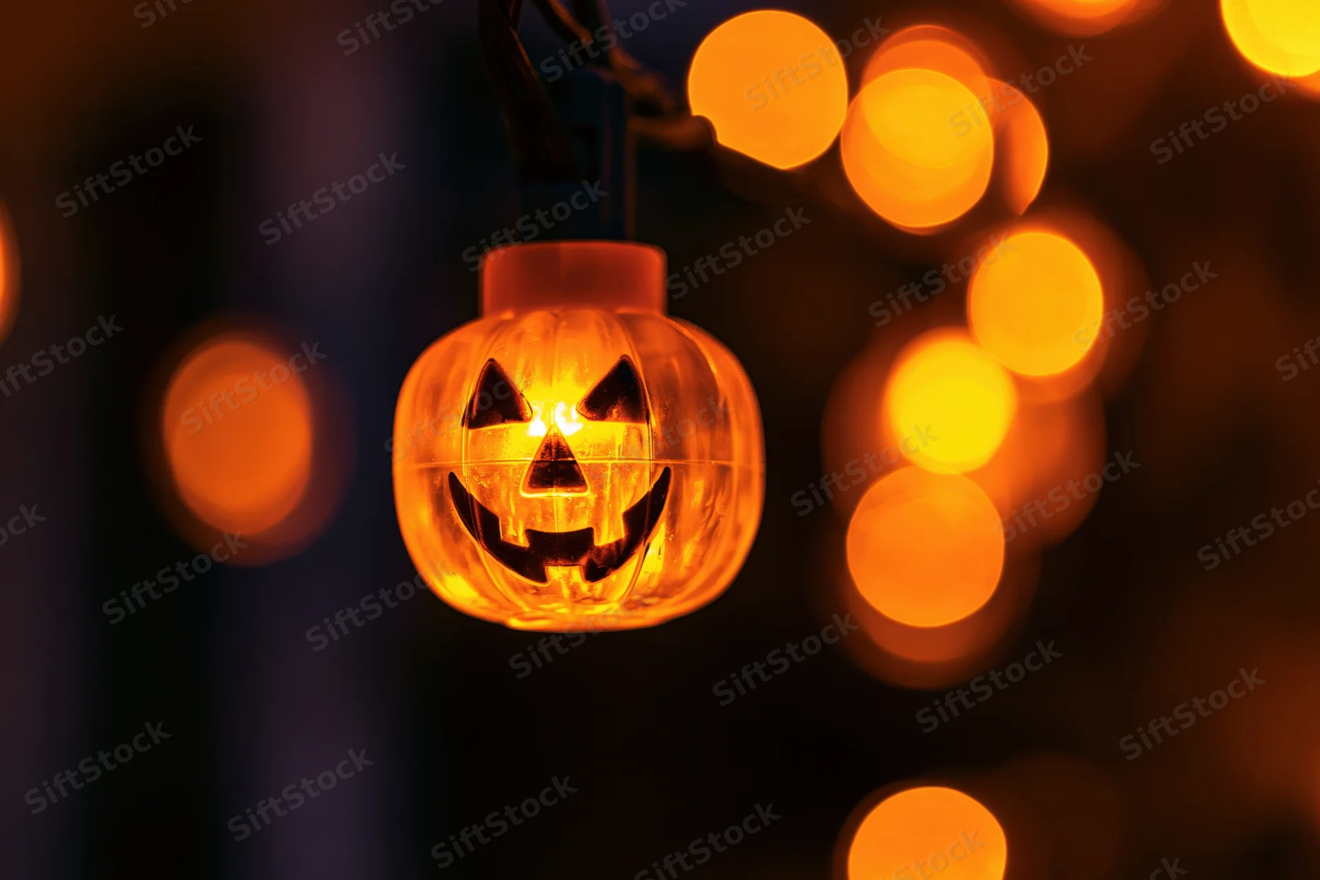 Glowing jack-o-lantern string light hanging in darkness with blurred orange lights in background