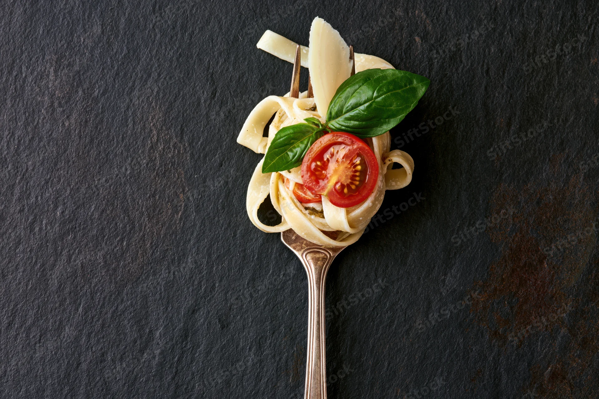 Vintage silver fork twirled with fettuccine pasta topped with tomato, basil, and parmesan cheese on dark slate surface
